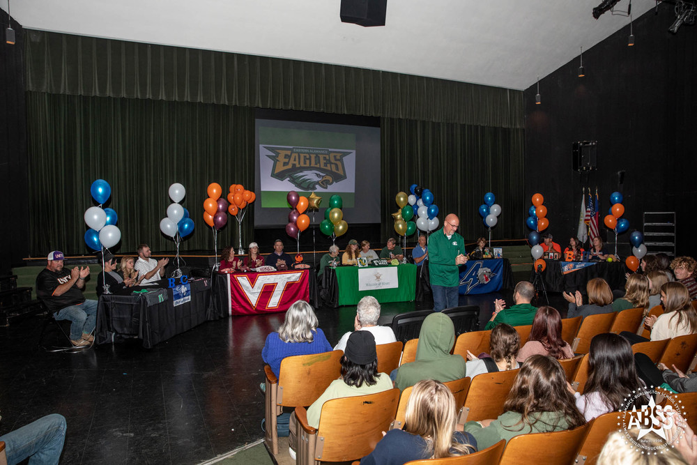 Student athletes seated at tables with their respective college banners in front and colorful balloons matching their future college colors