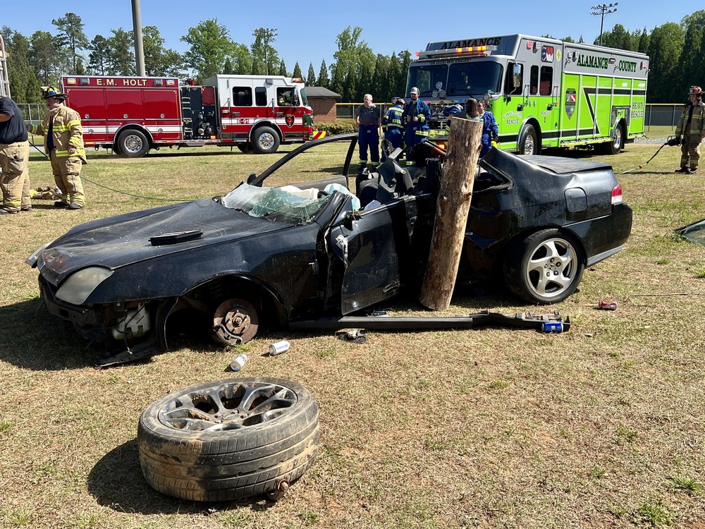 photo of wrecked car in mock crash.  tire on foreground.  left side of car hitting large tree trunk.  car top removed and windshield broken. Fire trucks in background.