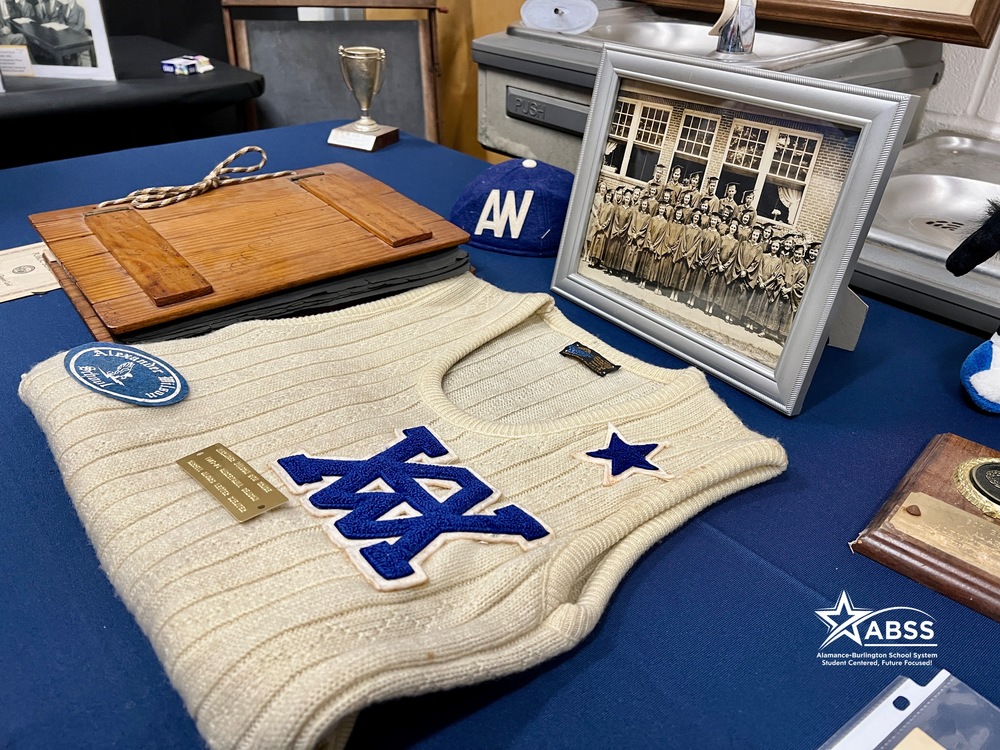 Photo of memorabilia on table.  Large AW on sweater with black and white photo of graduates in background