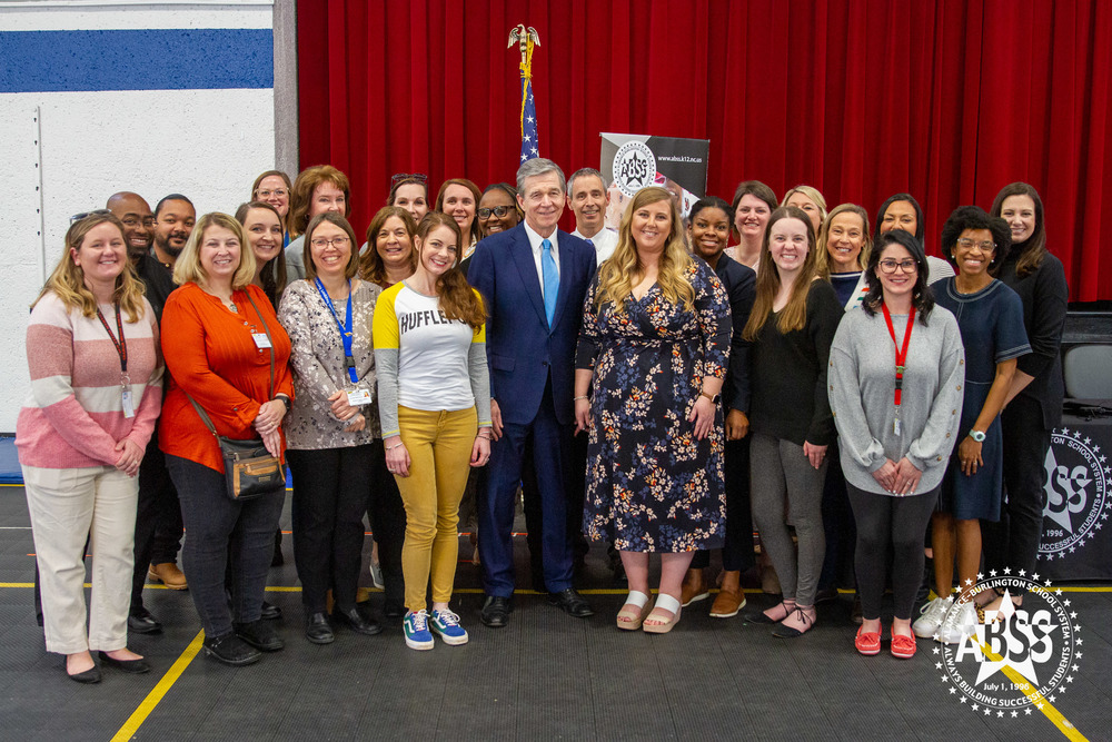 Photograph of Governor Roy Cooper standing with Alamance-Burlington school system counselors, and national Council of the year Meredith Draughn in the gymnasium at B Everett Jordan Elementary