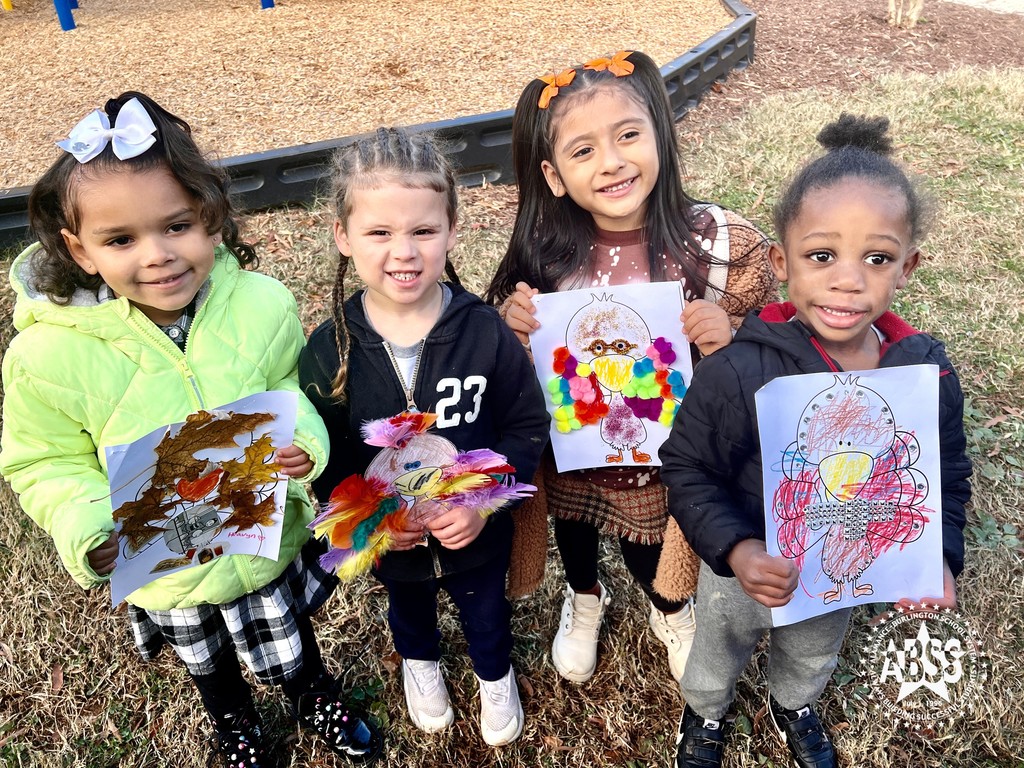 Pre-K students at Newlin Elementary standing outside near a playground holding turkey art they created and smiling at the camera.