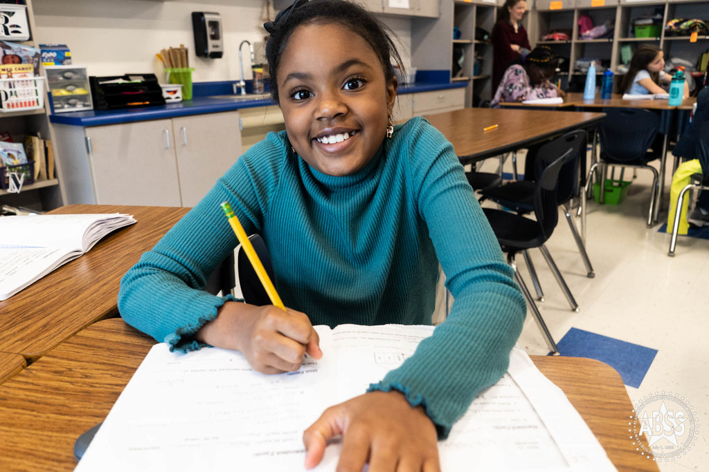 A young girl in a teal long sleeve shirt stops working in a workbook long enough to smile at the camera