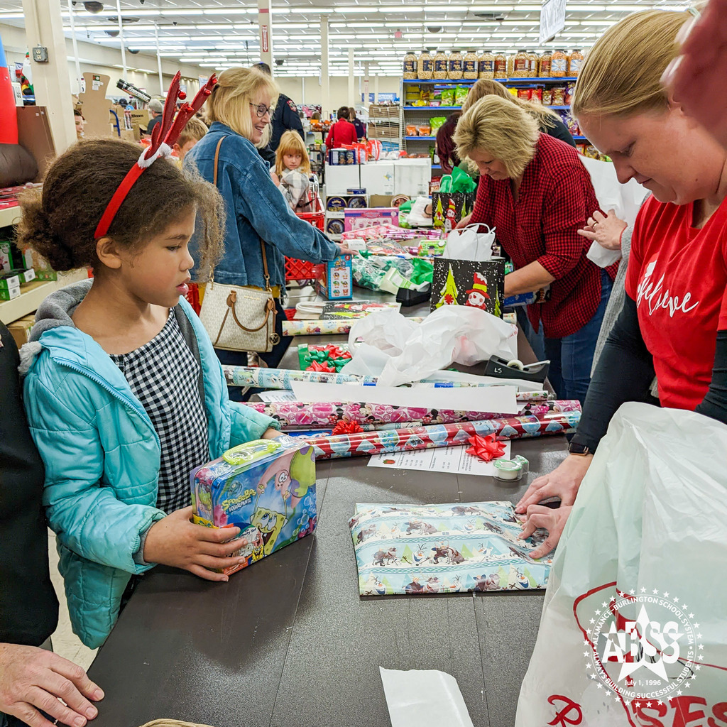 A young girl holds a toy at a gift wrapping table at Rose's in Burlington as volunteers finish wrapping another item in blue Frozen paper