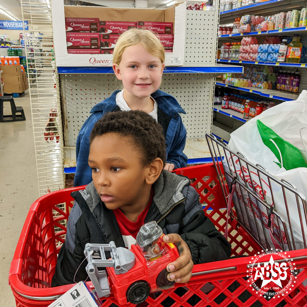 A young boy sits in a red shopping cart with his red fire truck toy while a student helper stands behind the cart and smiles at the camera