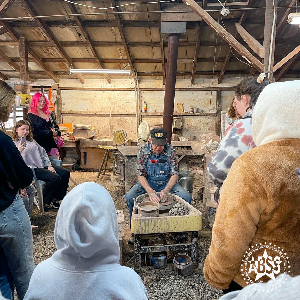 Students look on as a older, male potter in a red button down shirt and blue jean overalls demonstrates how to use your hands on a sculpting wheel to move clay