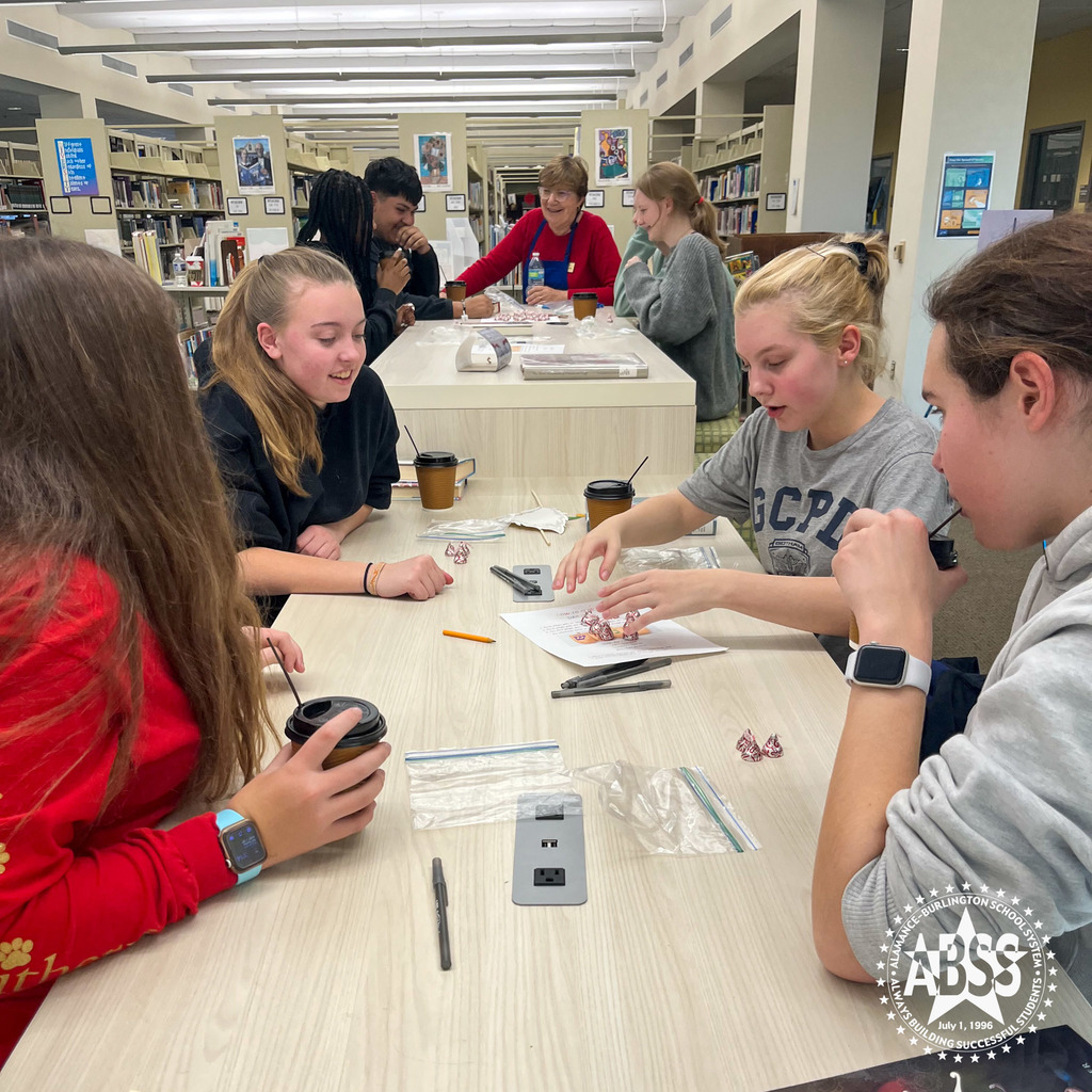 Four Early College students sit around a table in a library at Alamance County College discussing some books with coffee.  In the background are three more students talking to librarian Colleen Macklin.