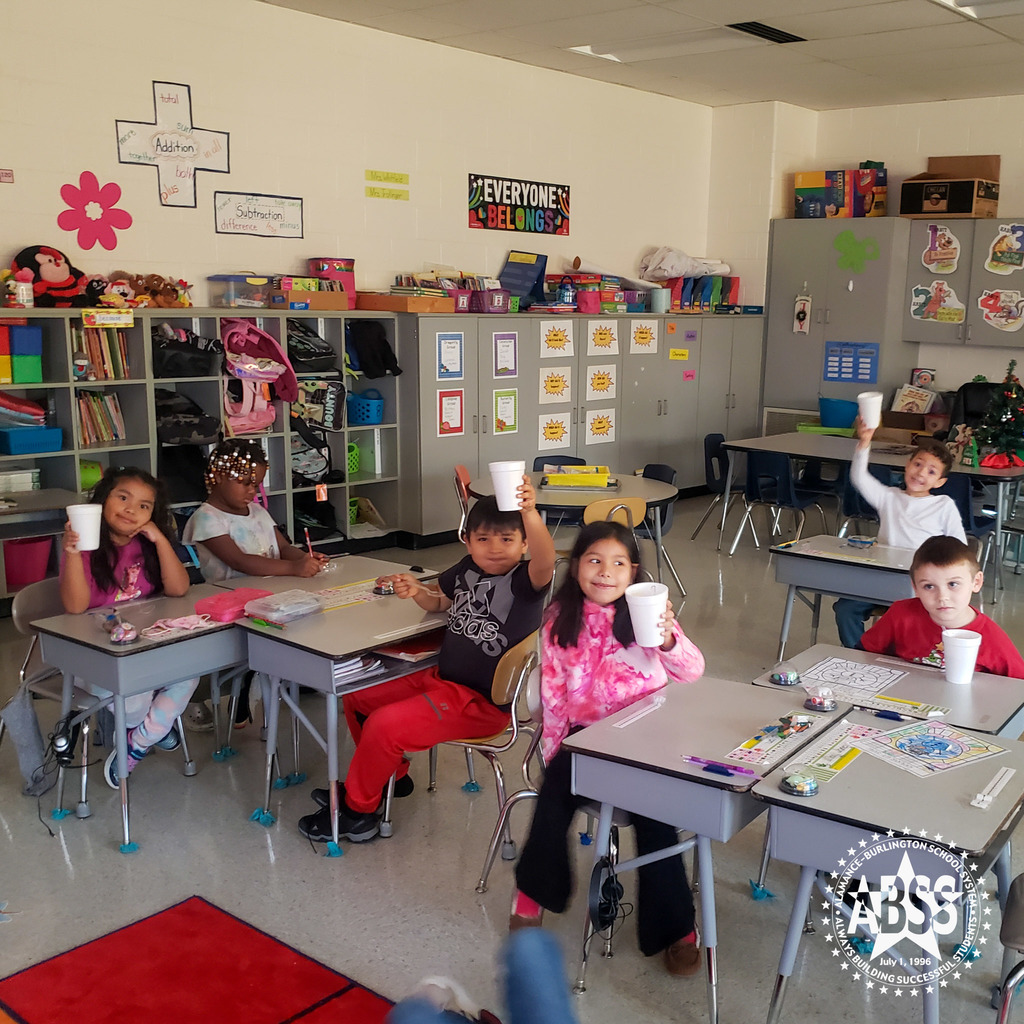 First graders at Hillcrest Elementary are sitting at their desks and four of the six are holding up a styrofoam cup of hot cocoa.  One girl is writing and the other is looking away with his cup sitting on the desk.