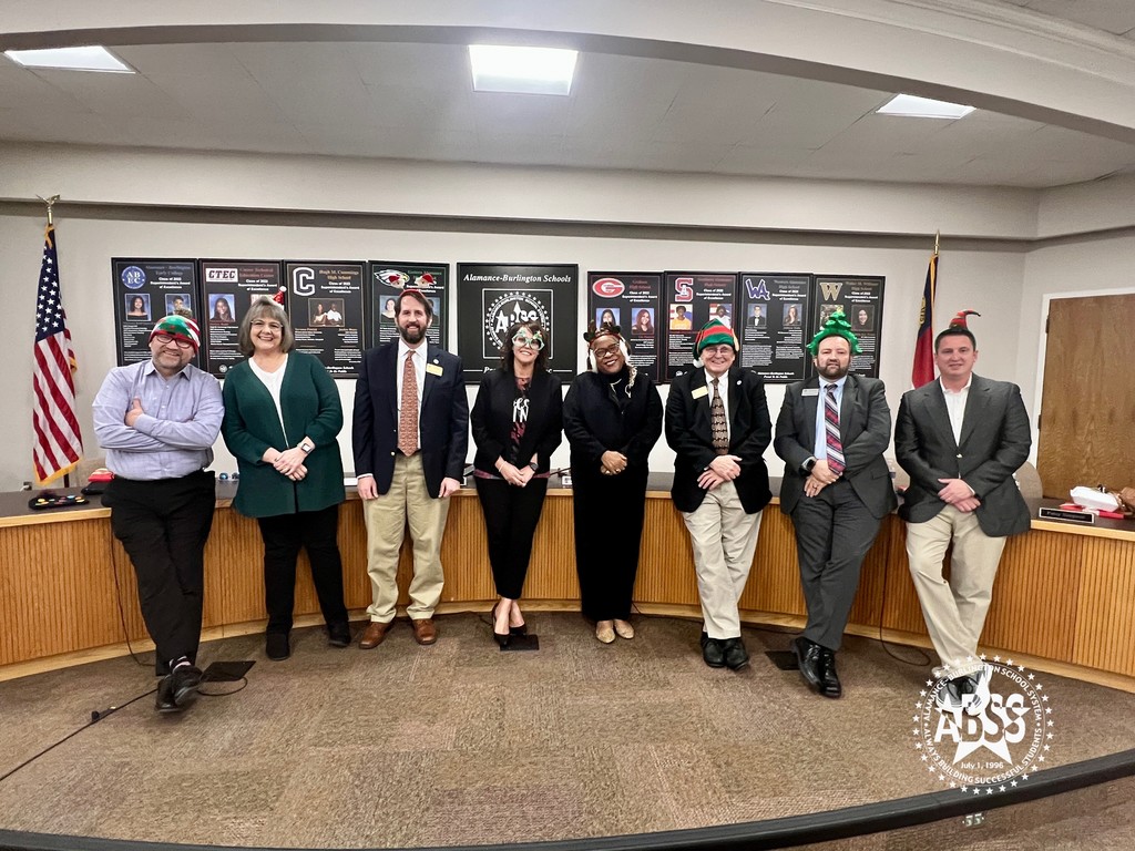 The Alamance-Burlington Board of Education and Superintendent Dr. Butler stand in the ABSS Central Office auditorium with festive holiday hats, standing side by side, smiling at the camera