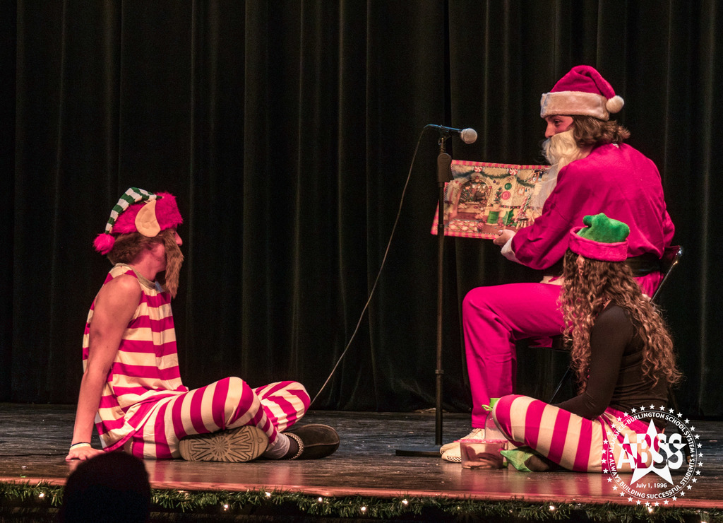 Three students in holiday garb on stage acting out a festive scene with a microphone