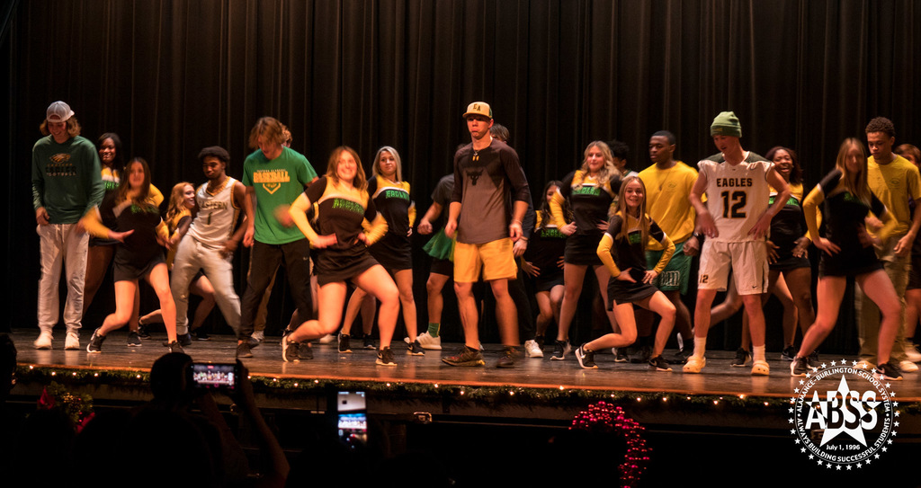 A combination of students in athletic gear, cheerleaders, and other students wearing green and yellow, dancing on stage.
