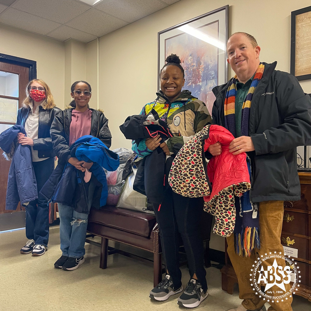 Patrick Vernon, teacher at Alamance Virtual School, two students, and another teacher, stand inside a building holding donated jackets.  On a couch behind them are more donated jackets in a large bag.