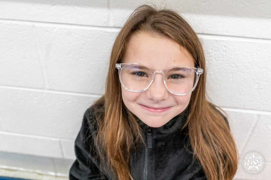 Elementary student with brown hair and clear glasses and wearing black jacket looks up and smiles at the camera in a school hallway