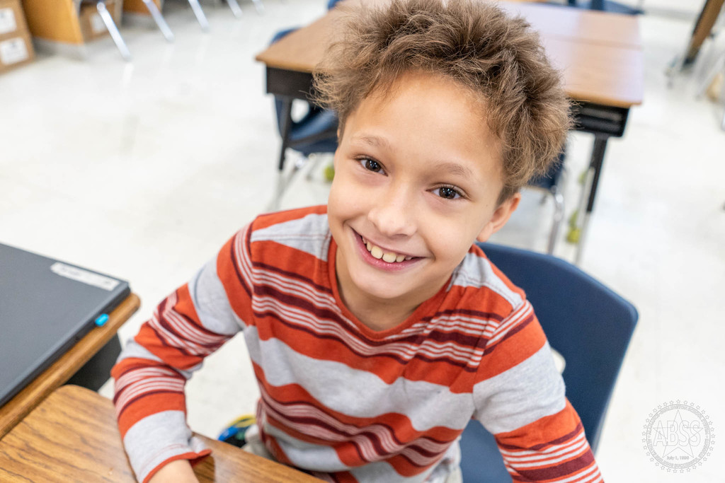 Elementary student with striped white, orange and red, long sleeve, shirt, and brown here looks up and smiles at the camera as he sits at his desk and works
