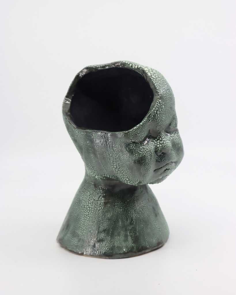 A pottery design titled: Ainsley Dial, Grade 11- Gold Key "Growth"  It is a green and black shaped baby head on a conical base.  The head has an opening on the top.