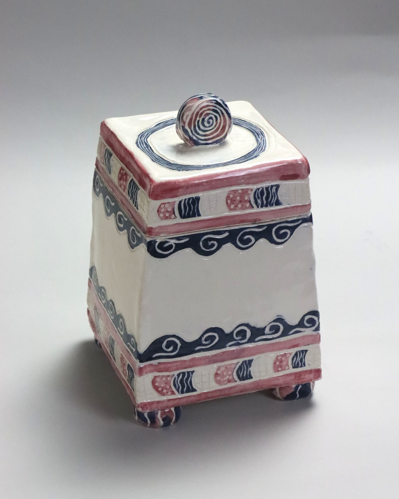 Pottery piece titled:  Kaitlyn Mason, Grade 10-Silver Key "Finding Atlantis".  It is a white rectangular prism with red and blue designs containing lines, waves, and arrows.  On the top is a solid circle protruding from the design surrounded by a blue swirl.