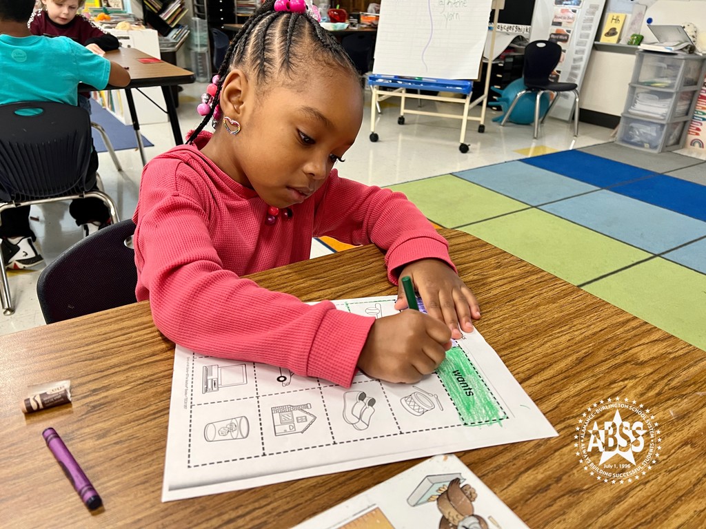 A kindergarten student uses a green crayon to color in the box with the text Wants on her activity paper