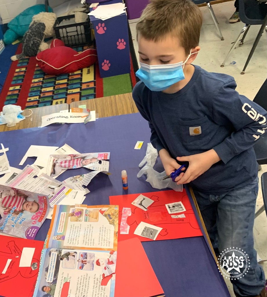 An elementary student wearing a surgical mask holds scissors and examines a magazine.  On the table in front of him are parts of the magazine he has already cut up.