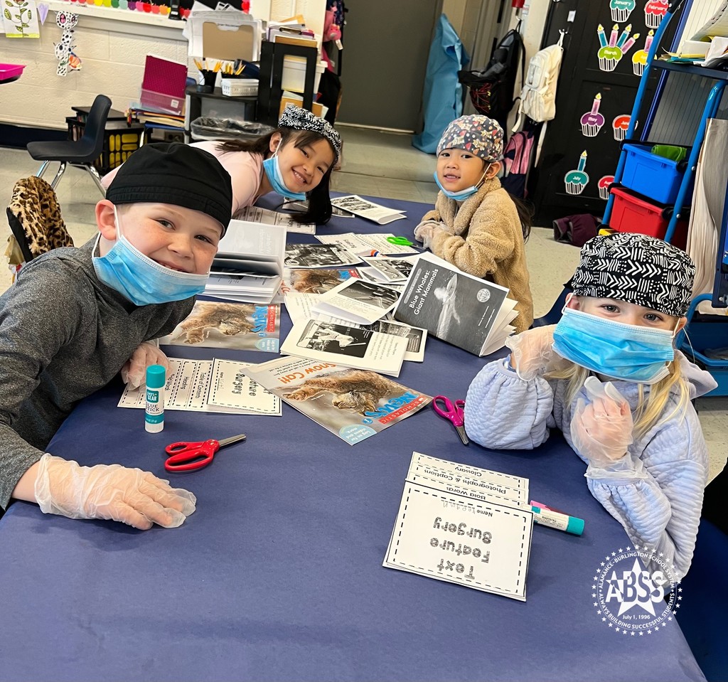 Four elementary students wearing surgical masks and knit caps are seated at a long table with paper booklets, magazines, and scissors in front of them.