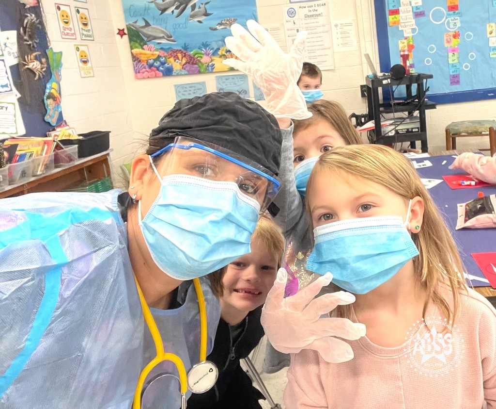 Nurse Kelly corm standing with three elementary students in a classroom. Two of the students in nurse quorum are wearing surgical masks and nurse. Quorum is also wearing protective goggles and a smock. There is one student looking underneath the other two students smiling without a surgical mask. In the background as another student standing with a surgical mask.
