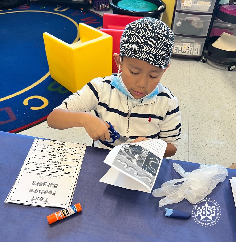 An elementary student is seated at a table and is using scissors to cut from his paper booklet. He is wearing a surgical mask when it is pulled down at his chin. He also has on a knit cap.  On the table beside him is a paper booklet that he is going to be gluing something into.