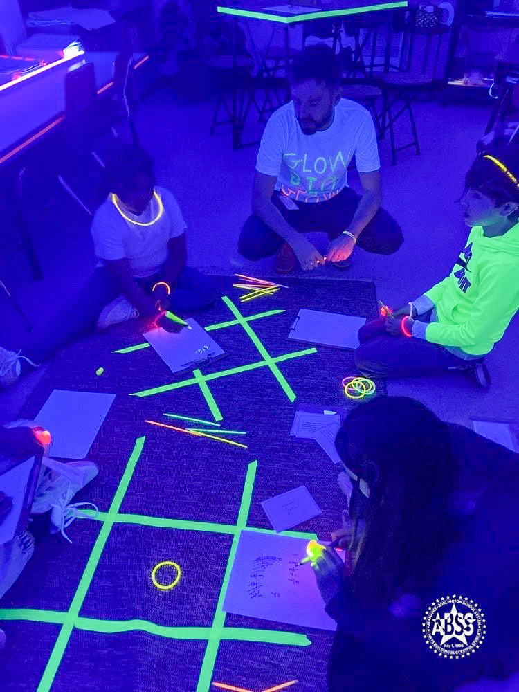 Teacher Mr. Hayes from Smith Elementary sits on the carpet around a big Tic Tac Toe board math activity made from tape.  Students are gathered around.  The classroom is dark and there is a blacklight which illuminates some colors in a neon glow.
