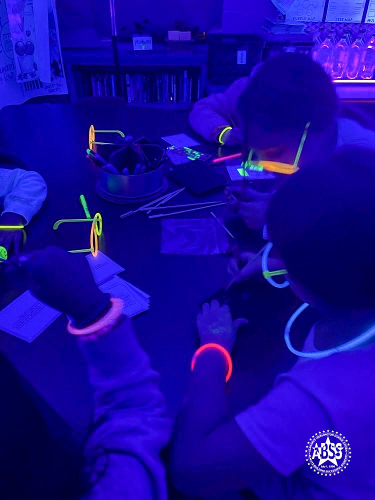 Students wearing neon glow bracelets and glasses work together around a table on a math activity.