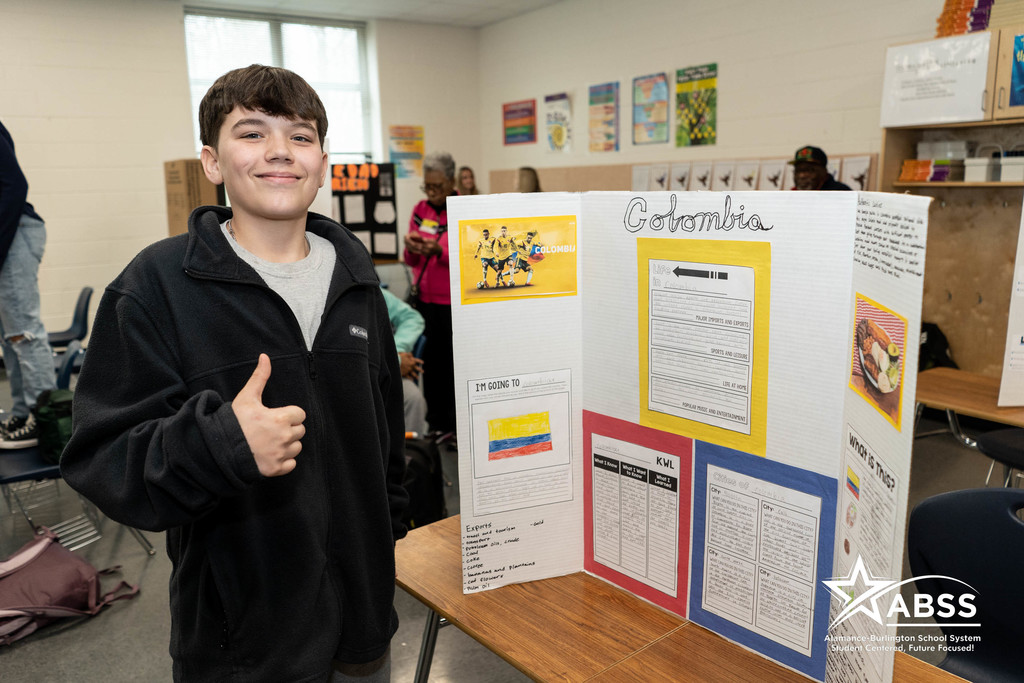A student at Western Middle School gives a thumbs up in front of his presentation board with information about Colombia