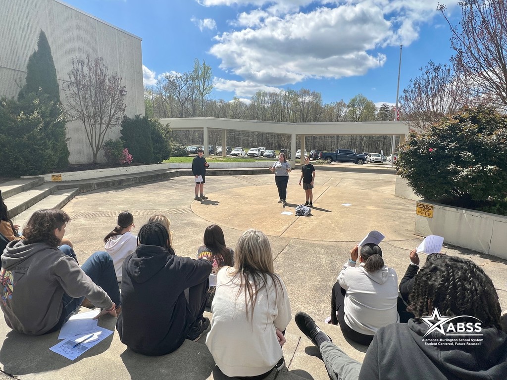A class of eight graders sits on concrete stairs around an outdoor concrete auditorium looking on at two students and instructor Stephanie Williams in the middle at Southern Middle School