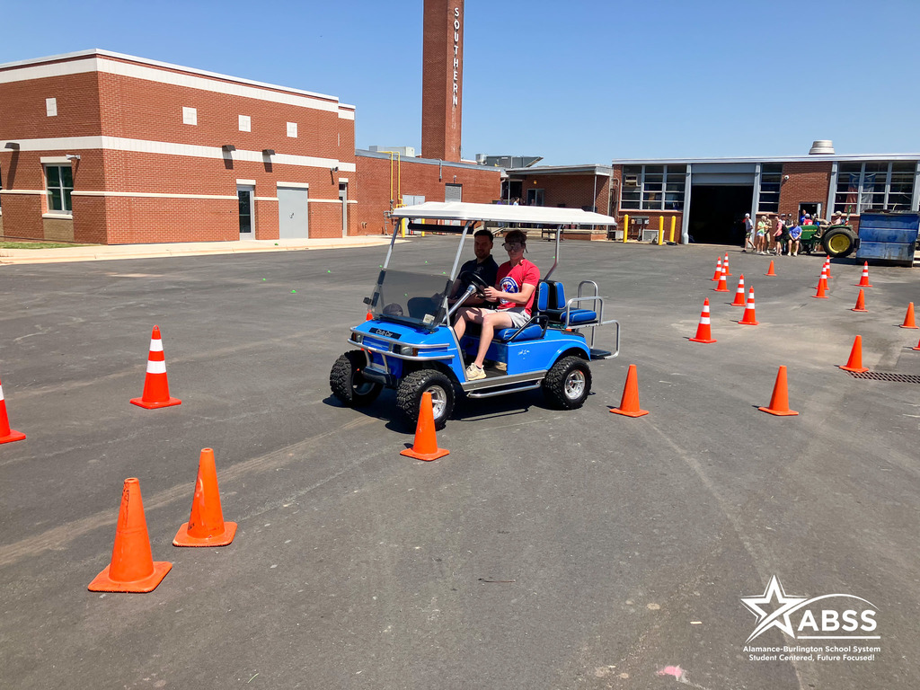 A student wears impaired vision goggles and drives a blue golf cart between cones under supervision of an officer