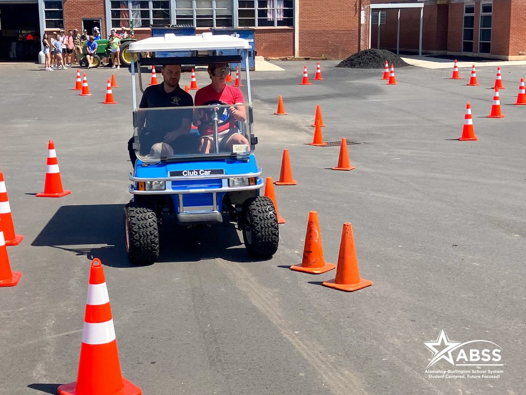 A student wears impaired vision goggles and drives a blue golf cart between cones under supervision of an officer