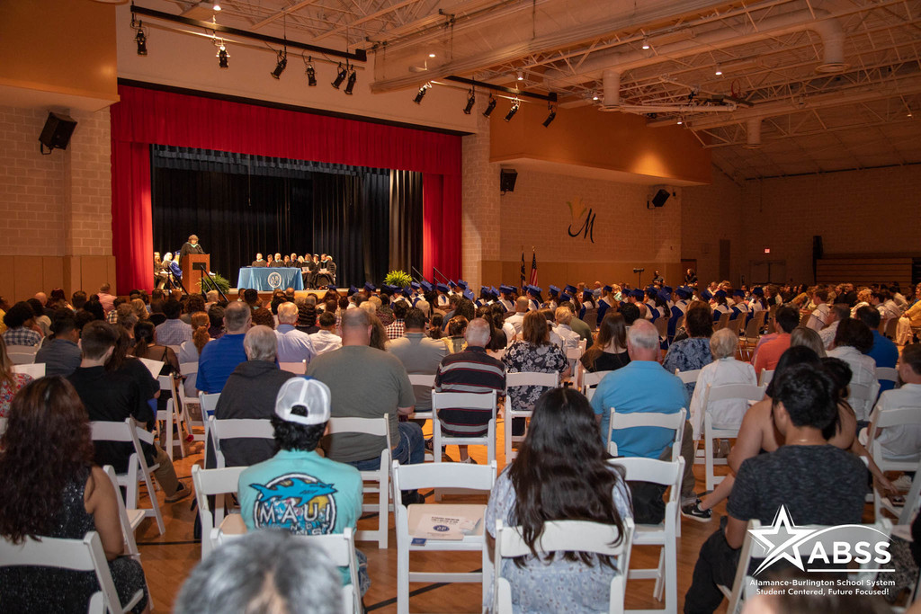 image showing crowd of guests at ABEC graduation. Facing stage showing graduates and ABSS Board on stage., Principal at podium.