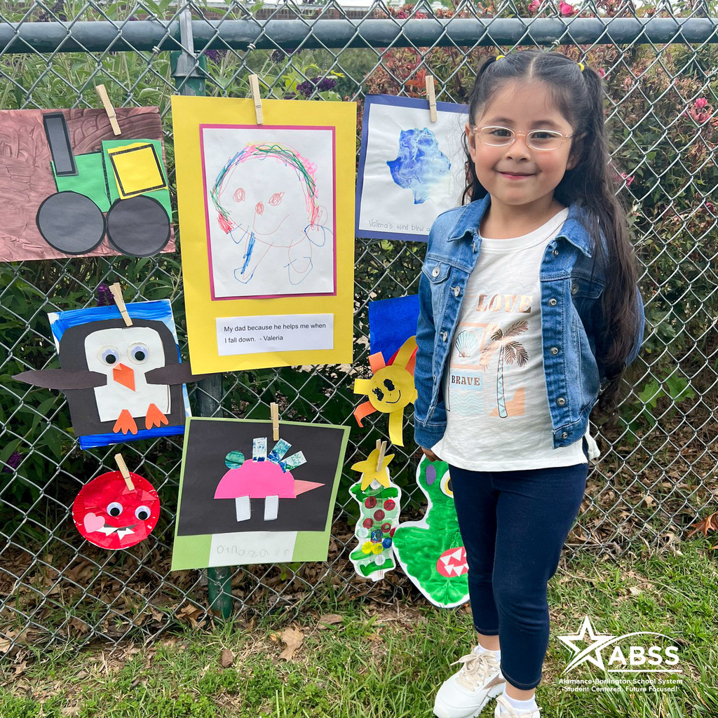 A Pre-K student at Smith Elementary stands in front of her artwork along a fence as part of an art walk