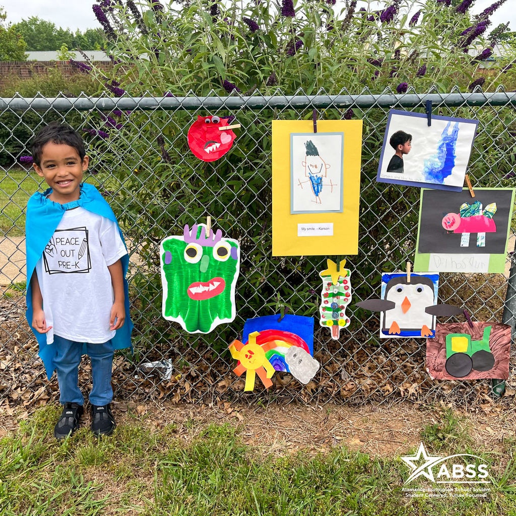 A Pre-K student at Smith Elementary stands in front of his artwork along a fence as part of an art walk