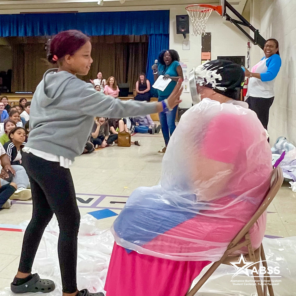 A student places a whipped cream pie in the face of a staff member wearing a large clear trash bag