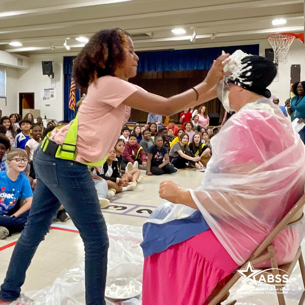 A student places a whipped cream pie in the face of a staff member wearing a large clear trash bag