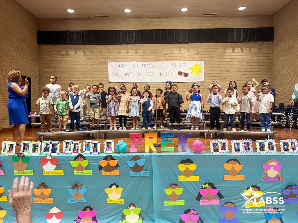 Pre-K students on a stage singing to the audience, in the foreground is a table with blue tablecloth and portraits of each student