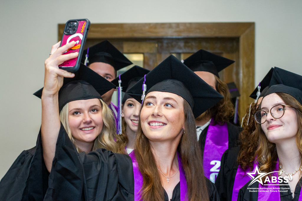 A group of Alamance virtual school seniors in cap and gown looking at a phone as one takes a large group selfie