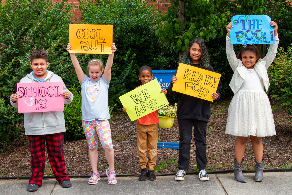 Five elementary students holding multicolored signs at various heights which spell out the phrase, "School is cool, but we are ready for the pool!"
