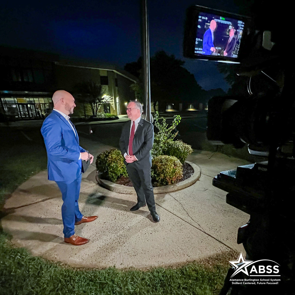 WFMY2 Anchor Ben Briscoe and ABSS Public Information Officer Les Atkins standing around a flagpole at Newlin Elementary for a live segment, lit by studio lights and a camera nearby is filming