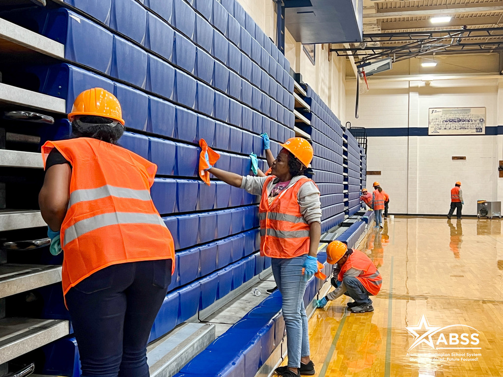 Workers in orange vests and hard hats wipe down the surface of bleachers in a gym