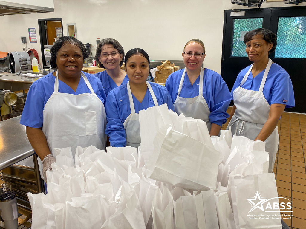 Team of school nutrition staff wearing scrubs and bibs standing behind a pile of white lunch bags