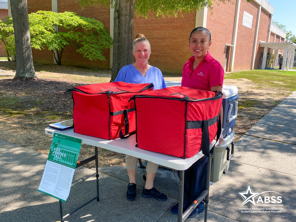 Two school nutrition staff stand behind red insulated bags filled with lunches for students