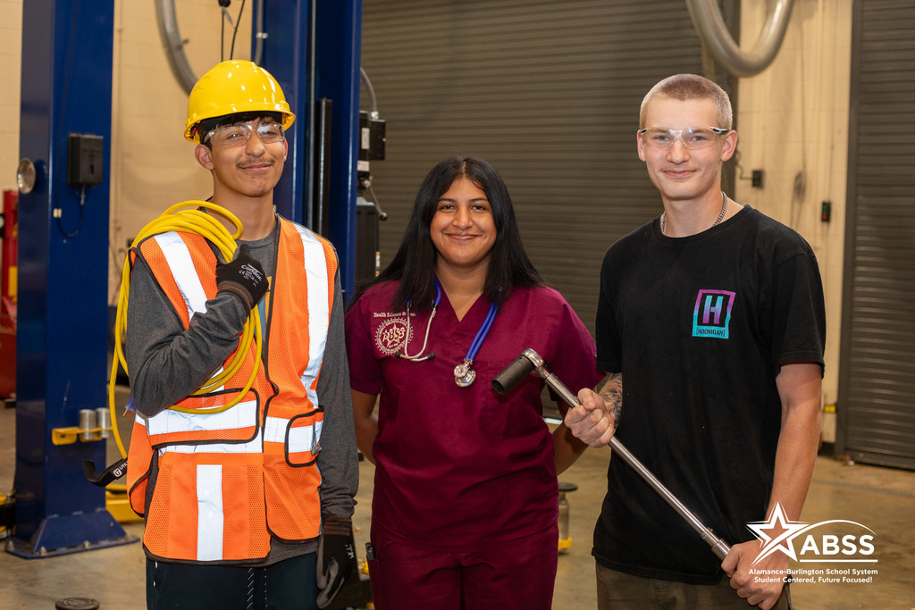 Three students in the Career and Technical Education program wearing a construction vest, nursing scrubs, and holding a tire iron