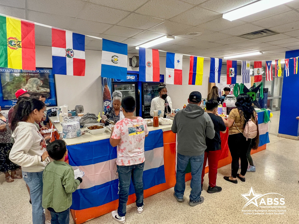 Families line up to try cultural foods in a buffet line lined with different flags and staff serving behind a long table