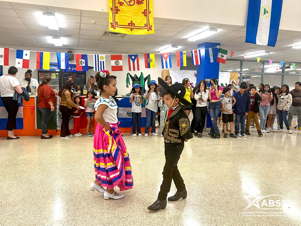 Two students in cultural Mexican attire dance in front of a line of families
