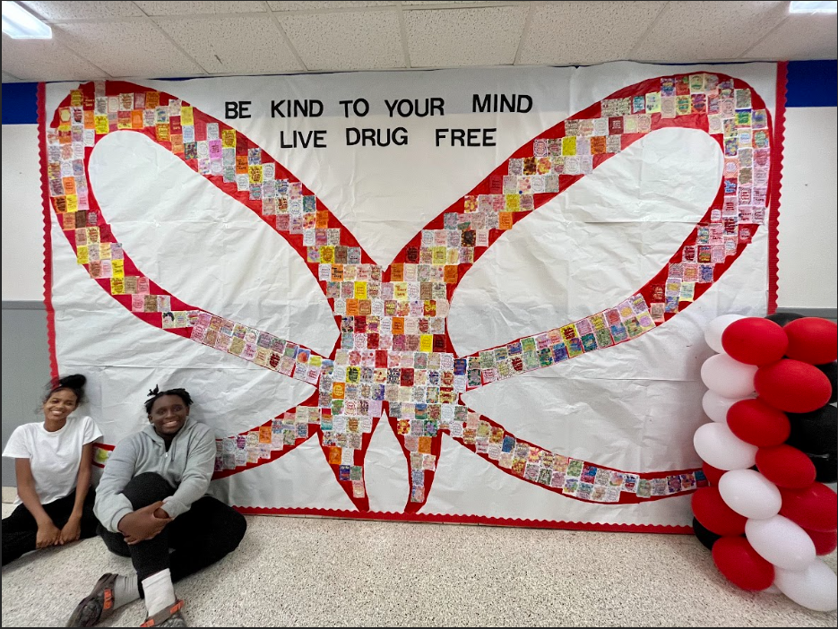 Two Broadview Middle school students sitting in front of a large Red Ribbon mural with the phrase Be Kind To Your Mind Live Drug Free