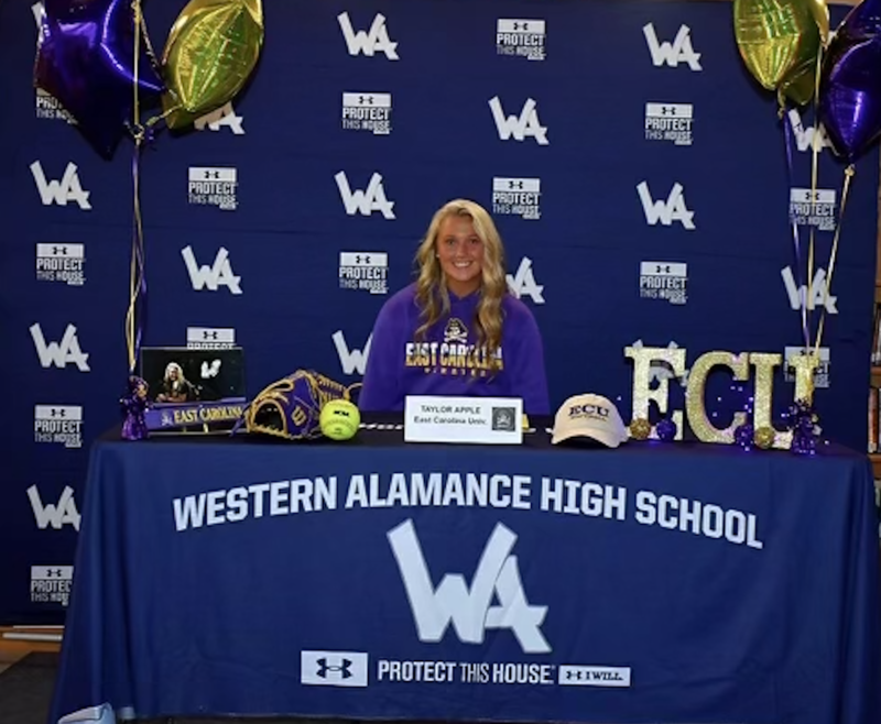 Western Alamance Senior Taylor Apple seated at table with ECU letters on it and softball ball and glove.  