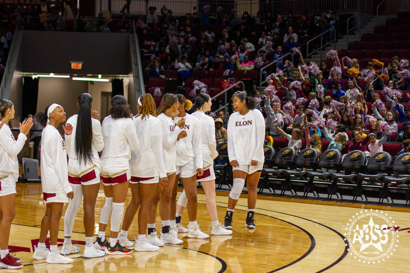 Elon University women's basketball players lined up on court pre-game while elementary students cheer in the crowd behind them.