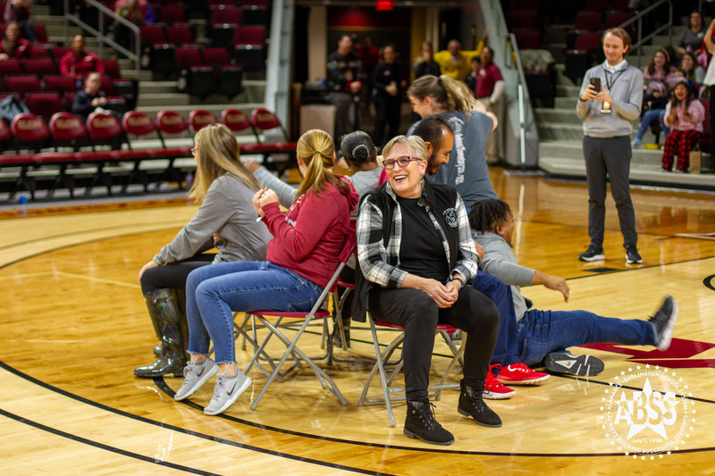 Five teachers are seated in a circle on the basketball court during a game of musical chairs.  One has fallen down after not getting a seat in time.