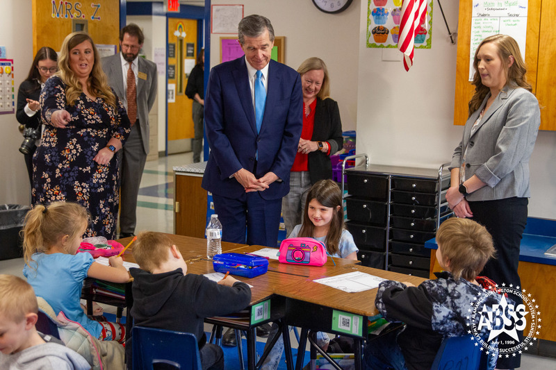 Governor Roy Cooper stands beside a second grade student in a classroom with teacher to his left and principal behind him, at B Everett Jordan elementary